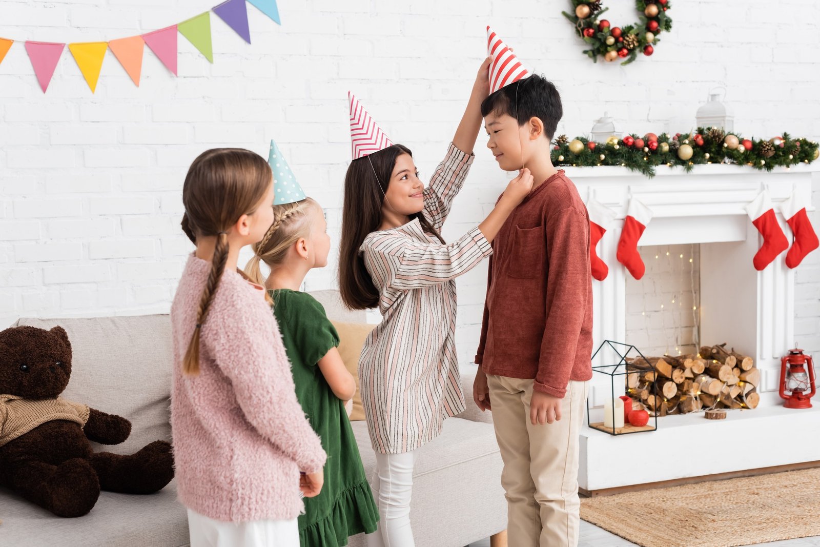 Smiling girl wearing party cap on asian friend during birthday party and christmas celebration